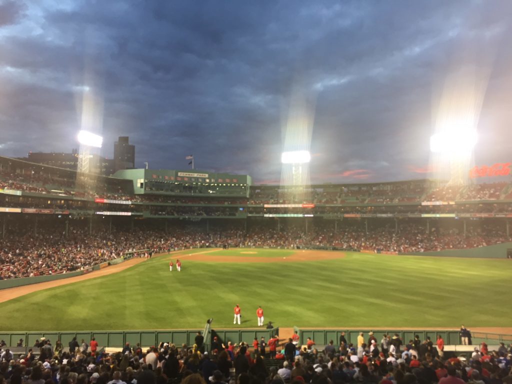 Cloudy skies, bright lights and a grassy baseball field with people below