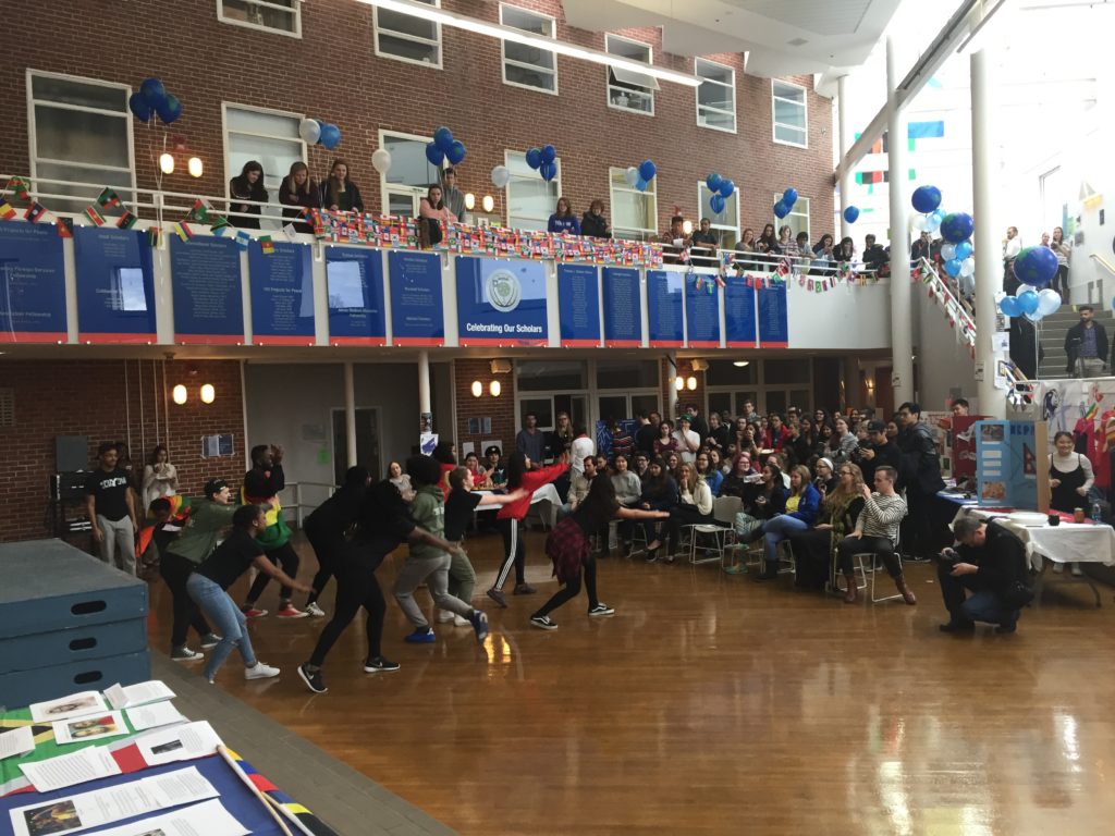 There are students sitting in chairs and standing above watching a student group do a dance performance