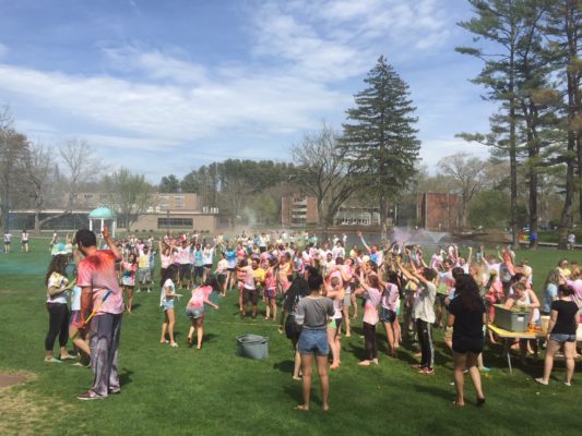 A group of students in wite t-shirts throwing powder at each other in a grassy field with blue skies