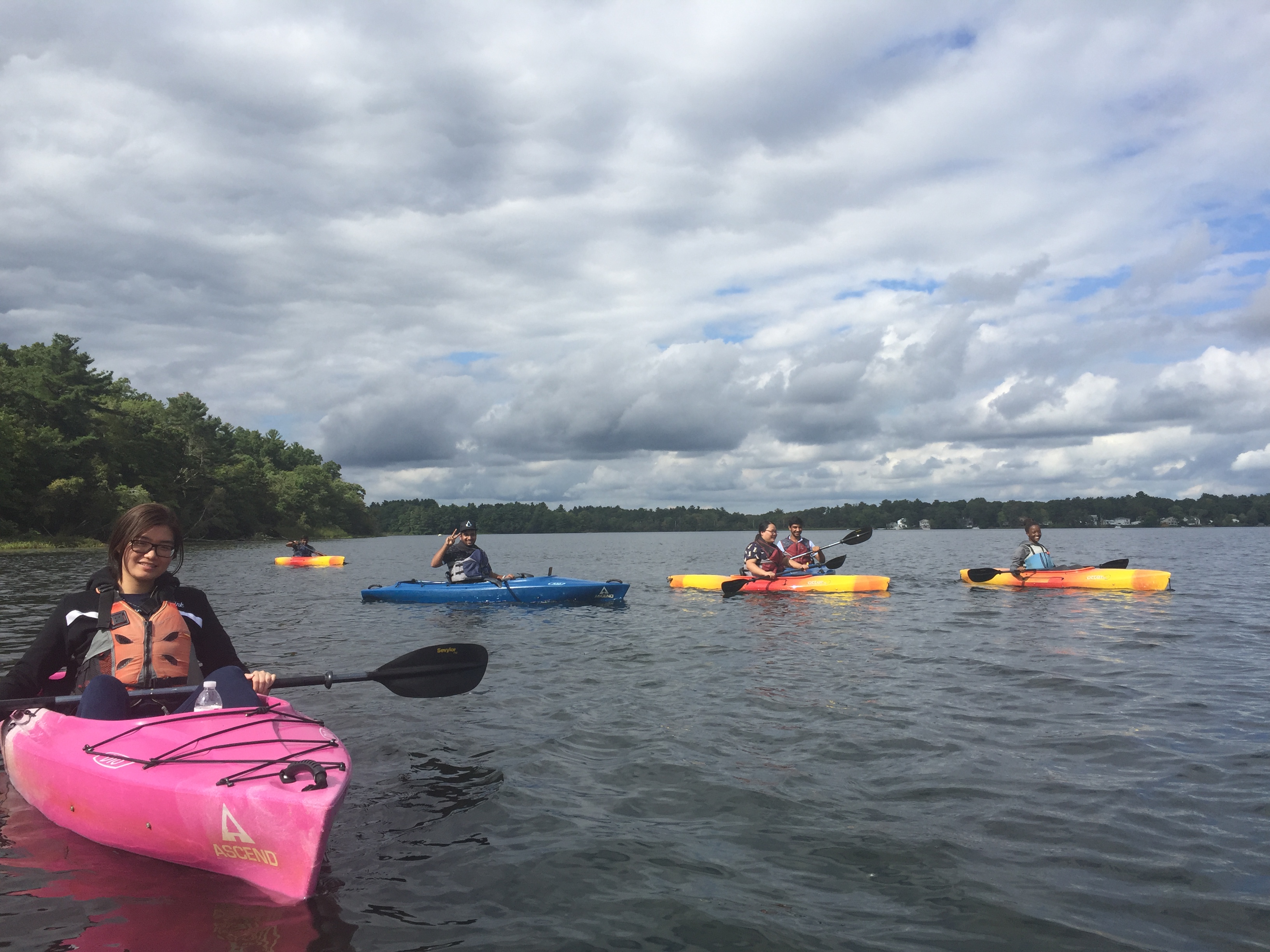 This is a group photo of six students in the water kayaking with clouds in the sky.