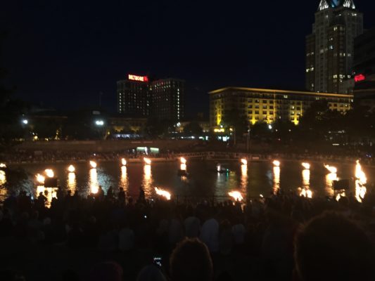 A photo of several firepits in the middle of a river in Providence. There are spectators sitting and standing nearby