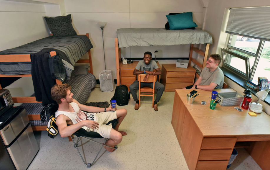 Here is a photo of a triple bedroom. There are two bunk beds with another bunk bed and a desk underneath it. There are three people sitting around in chairs in a circle. 