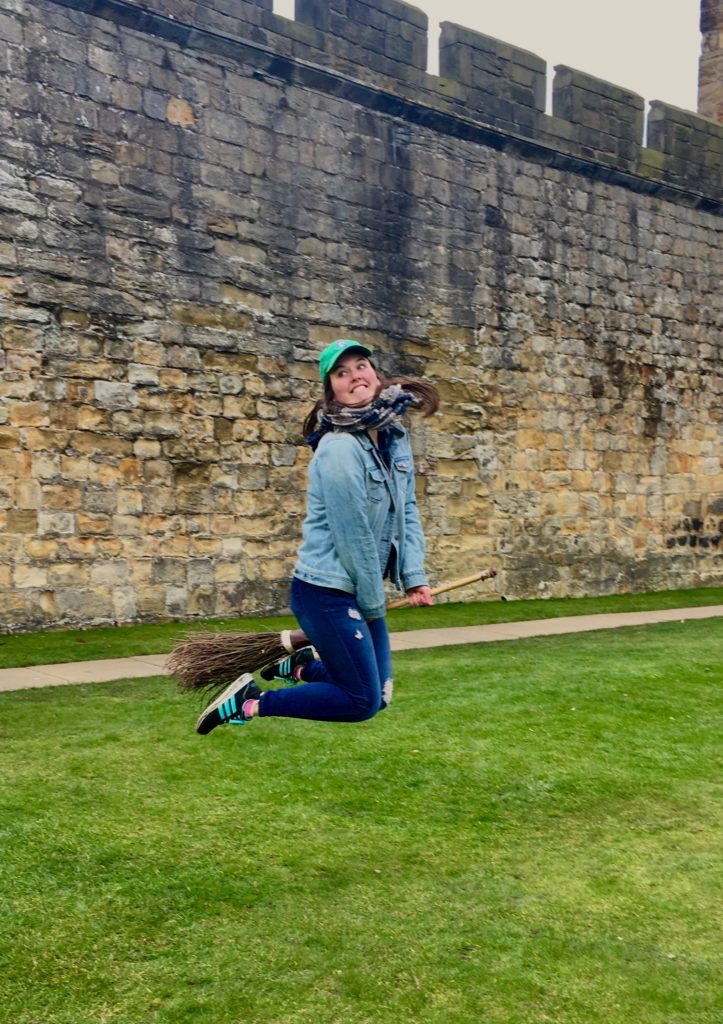 a photo of a woman jumping in the air on a broom. She is standing on grass with a castle wall behind her.