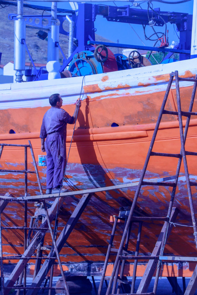A photo of a man standing on scaffolding while painting a boat red