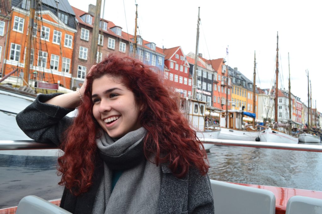 A photo of a woman with red hair smiling on a boat. There are multiple different colored houses behind her.
