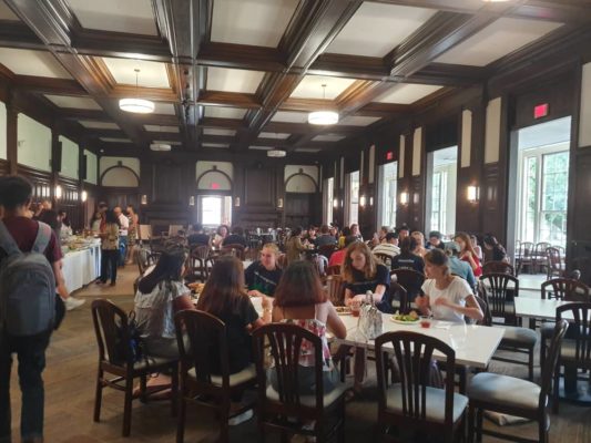 A group of people sitting around numerous tables eating food on wooden square tables