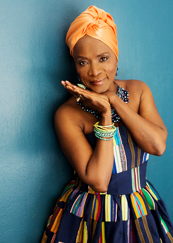 artist standing against a blue background wearing an orange hair wrap and a colorful dress, her hands are together and resting under her chin
