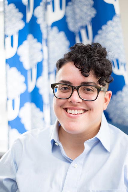 Aimi Hamraie, an olive-skinned Iranian person with short dark curly hair, smiles at the camera. They wear rectangular glasses and a blue button-up shirt. Behind them is a blurry blue background of blue curtains with white trees on them.