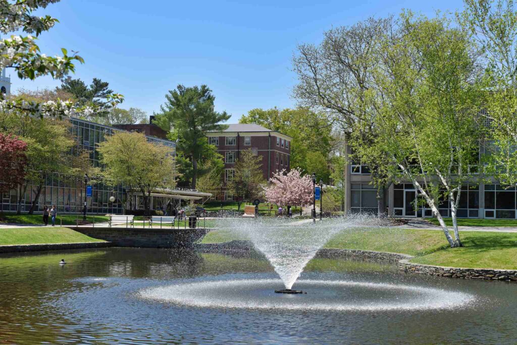 A pond with a fountain in the middle, with buildings and trees in the background.