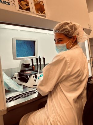 A student in a lab coat, mask, and hair net smiling in front of a microscope.