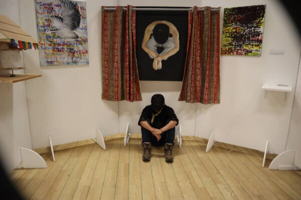 A person sitting on the ground against a wall with their head bowed.