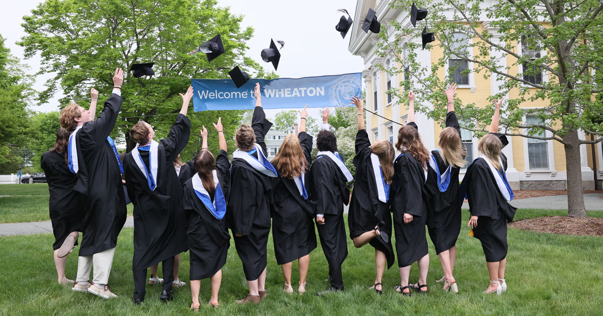 Graduates toss their caps up in front of Welcome to Wheaton sign