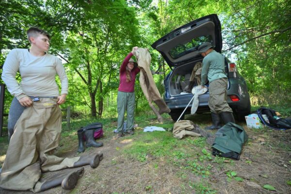 Three individuals don hip waders and long-sleeved shirts, long pants and hats outside a car parked near woods.