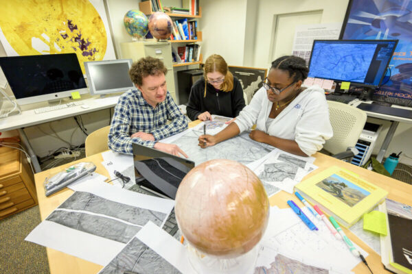 Professor Geoff Collins works with two students sitting at a desk.