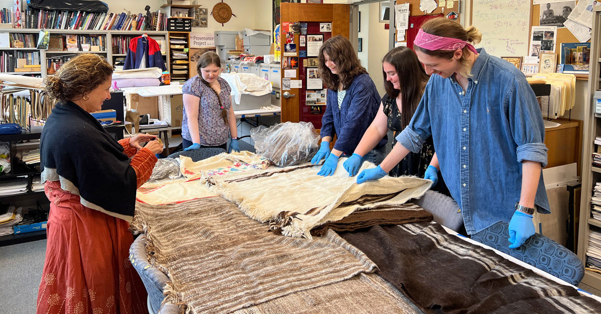 Five individuals inspect a tapestry resting on a table.