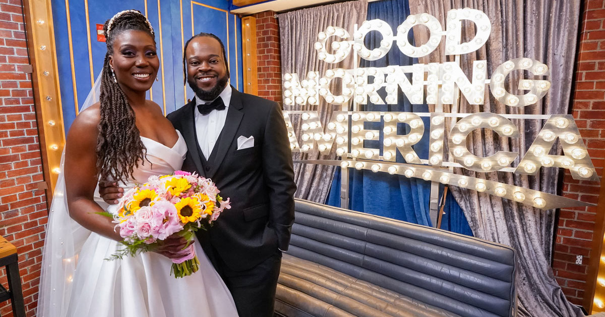 A woman in a wedding dress holding a bouquet of flowers stands beside a man in a tuxedo next to a Good Morning America sign.