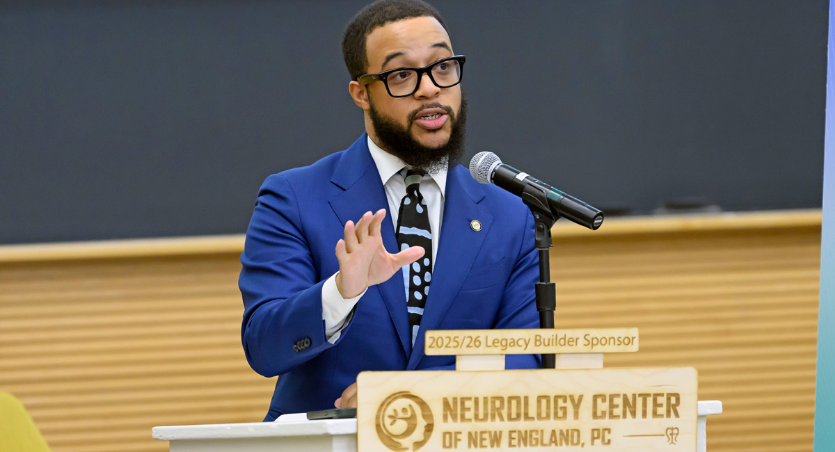 A man with short hair, a mustache and a beard, wearing glasses and a royal blue suit, speaks from a podium in front of a sign that reads "Neurology Center of New England, PC"