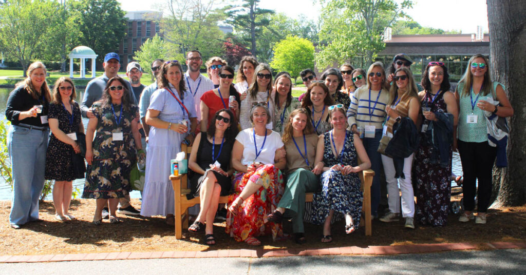 A group of 27 individuals are out side on a sunny spring day, looking at the photographer, with green trees and buildings in the distance behind them.