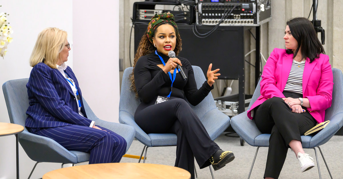A Black woman wearing a black shirt and pants speaks via a hand held microphone while seated. A woman wearing a blue suit is seating on the left and a woman wearing a pink blazer and black pants is seated on the right.