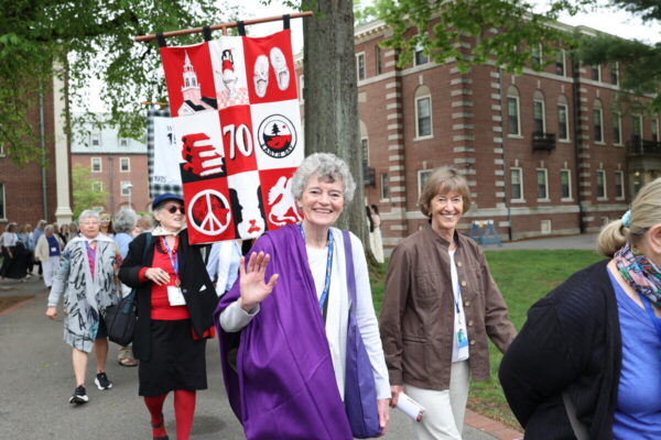 A group of women smiling and one waving. One woman has curly gray hair and is wearing a purple garment, while the other has short brown hair and is dressed in a brown jacket. Behind them, a banner featuring various symbols, including peace signs and numbers, is being carried.