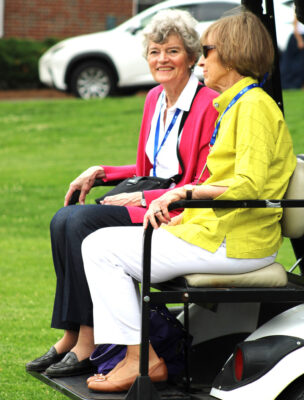 Two women sit on the back of a golf cart. One is wearing a pink coat, white shirt and dark pants. The other is wearing a yellow shirt and white pants.