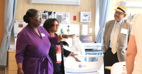 Three individuals in a hospital room. Two women on the left face a man on the right. One woman is wearing glasses and a purple dress, the other has glasses, a black coat, salmon shirt and dark pants. The man is wearing a beige sportcoat, hat and tie, white shirt and dark pants.