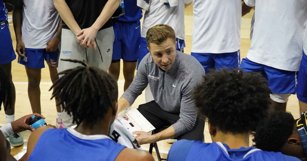 An individual with short hair wearing a grey sweatshirt speaks with a huddled group of basketball players.