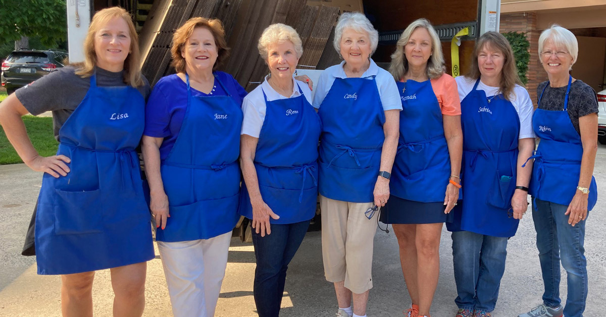 Seven women, each wearing a blue apron over their clothes, stand in front of a moving truck.