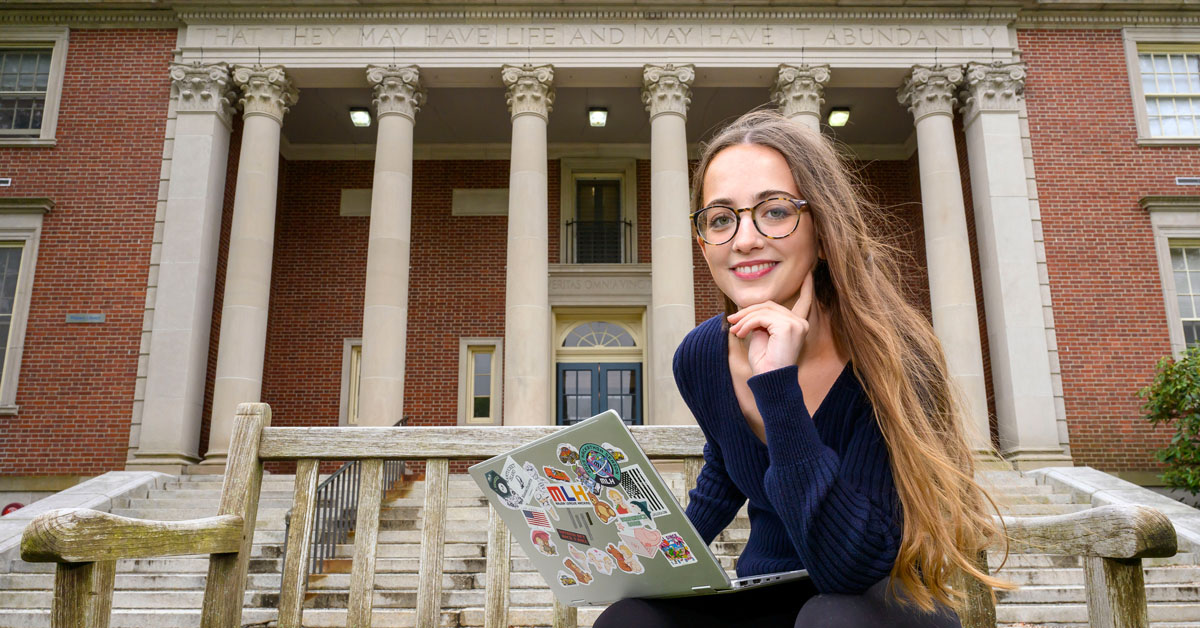 A woman with long brown hair wearing glasses, a dark blue shirt and black pants sits with an open laptop in her lap in front of a brick building with large columns.
