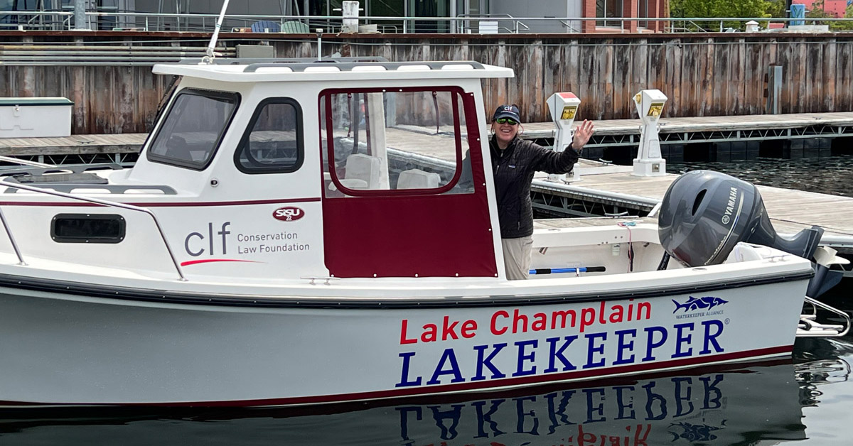 An individual wearing a dark coat, light pants and dark baseball cap waves from a boat in the water at a marina. The side of the boat reads Lake Champlain lakekeeper.