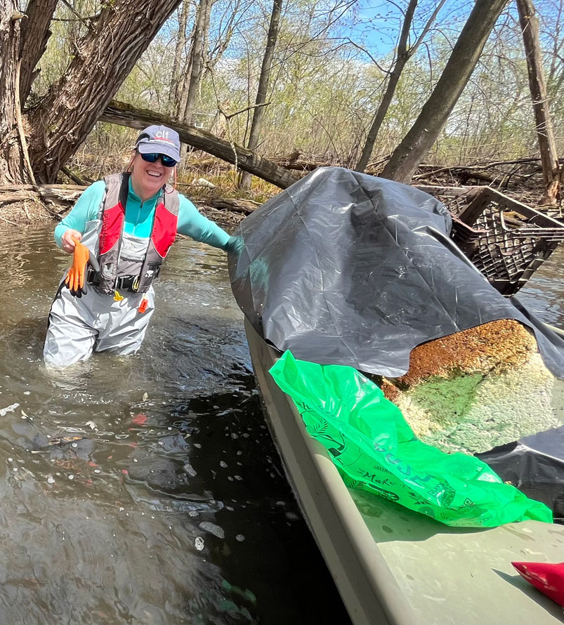 A woman wearing hip waders, a blue shirt, a baseball cap and sunglasses stands in water up to her thighs beside a boat carrying a tarp, a foam block, a plastic bag and an egg crate.