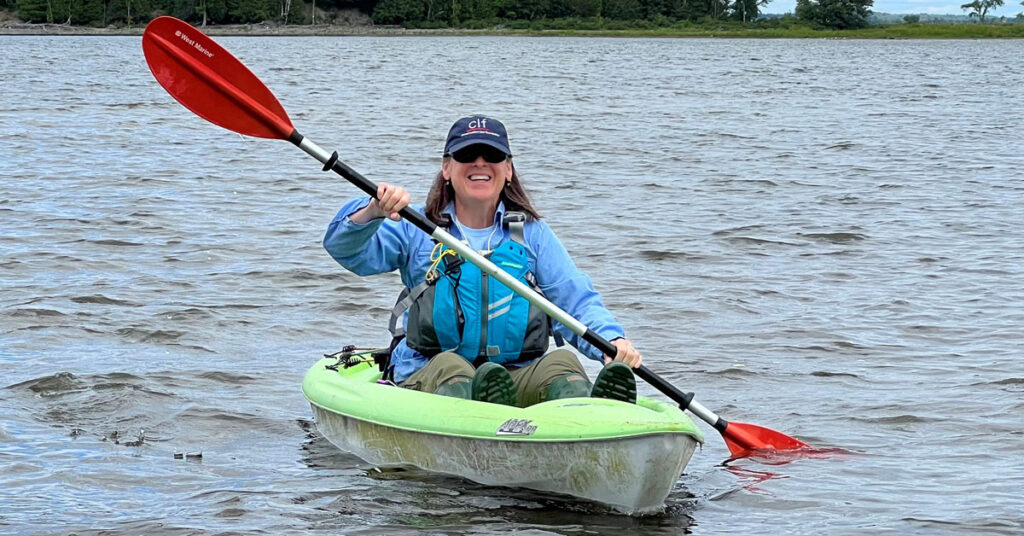A woman wearing a blue jacket and baseball cap and sunglasses paddles a light green kayak.