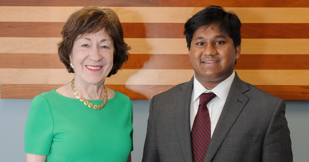 A smiling woman with short brown hair wearing a green dress stands next to a young smiling Black man with black hair wearing a grey suit, white shirt and red tie.