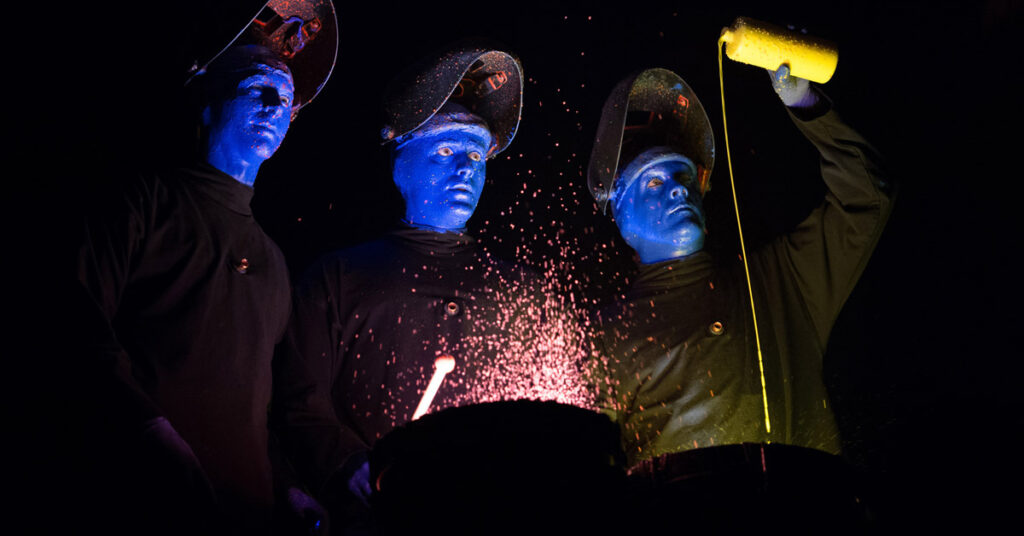 Three individuals with blue painted faces wearing black shirts stand in front of drums as one individual pours yellow paint into a drum.