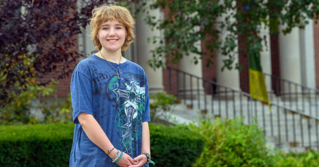 A smiling young woman with wavy light brown hair wearing a blue patterned shirt stands in front of a brick building with large white columns and green bushes.