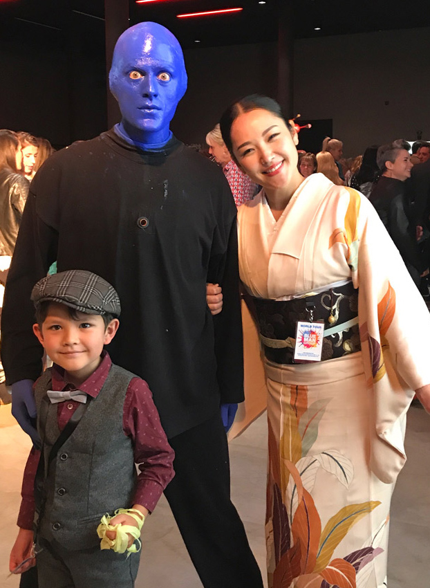 A man with a blue painted face wearing a black shirt and black pants stands beside a smiling Asian woman with black hair wearing a beige robe with a floral pattern. A young boy wearing a gray derby cap, vest and pants with a maroon shirt and light gray bow tie is standing in front of the man.