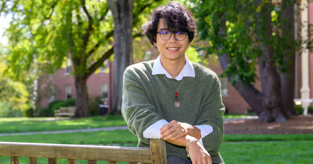 A smiling individual with wavy dark hair wearing glasses a while collared shirt with a faded green sweater over it leans on the back of a wooden bench. Trees with green leaves and a brick building is in the background.