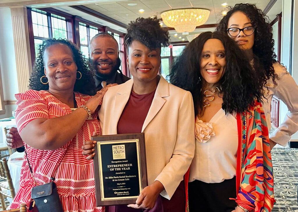 Five smiling Black individuals wearing business attire. A woman in the middle, wearing a red shirt and white sportcoat holds a plaque that reads "Entrepreneur of the Year"