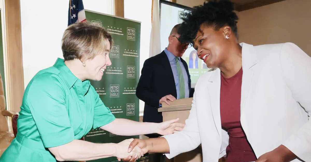 A smiling woman with brown hair wearing a green dress shakes the hand of a smiling Black woman with black curly hair wearing a red shirt and white sportcoat.