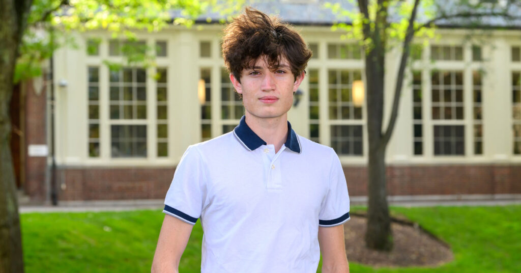 A young man with wavy dark hair wearing a white t-shirt with a blue collar stands outside in front of a trees with green leaves and a brick building with large paneled windows.