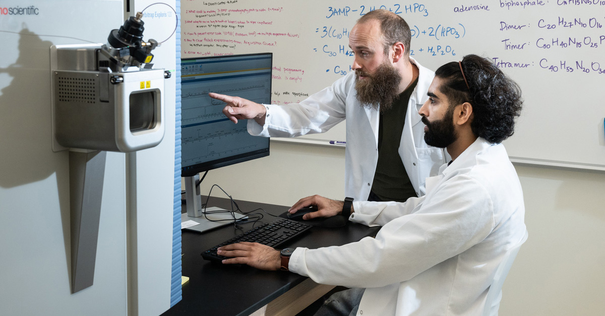 A man with thinning hair, a mustache and beard wearing a white lab coat and a dark shirt points at a computer monitor while a younger man with dark wavy hair, a mustache and beard wearing a white lab coat types at the computer's keyboard.