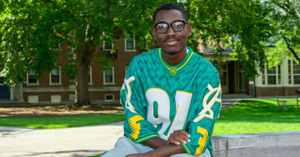 A smiling Black man with short black hair wearing glasses and a teal-colored jersey sits outside on a stone wall. Trees with green leaves and brick buildings are in the background.