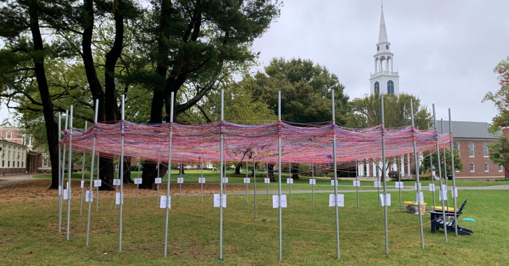 A collaborative art project of colorful skeins of yarn tied to a central metal pole, and stretched around several poles on a circular perimeter. The project is located on green grass with trees and a church with a white steeple in the background. 