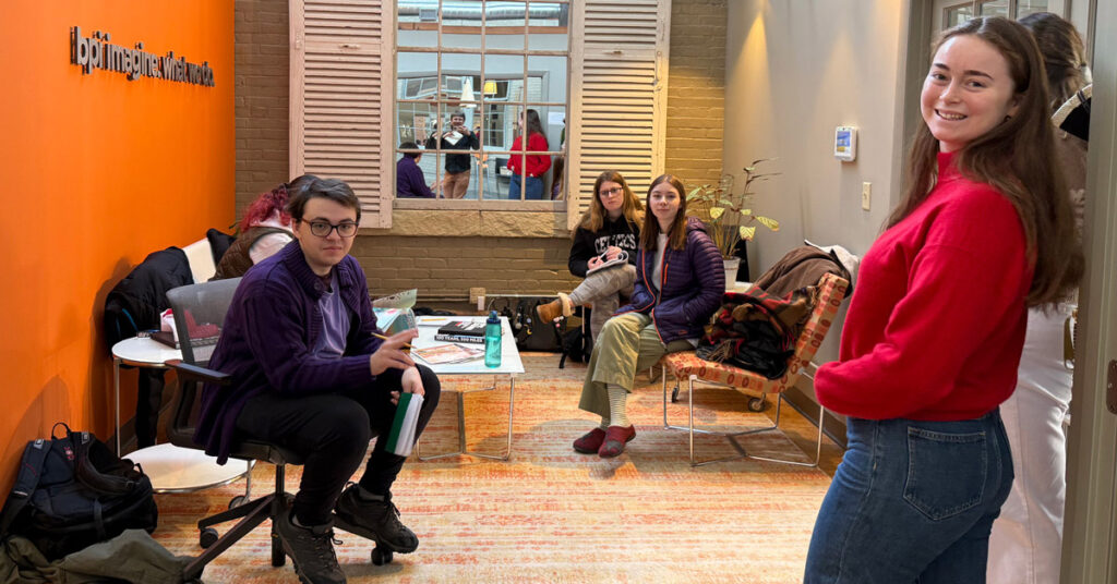 A smiling young woman with dark hair wearing a red sweater stands in a room while four young men and women sit.