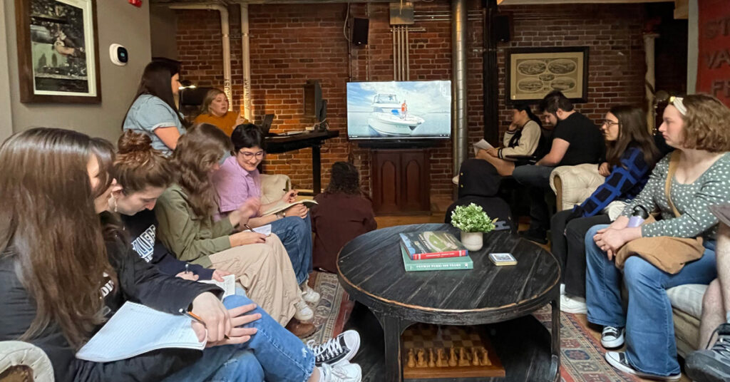 A group of college-aged students sit on furniture in a small room with some art on the walls and a small round coffee table between them.