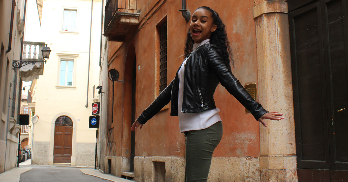 A smiling young woman with dark hair wearing a white shirt, dark coat and pants stands on a narrow street between buildings with orange stucco walls.