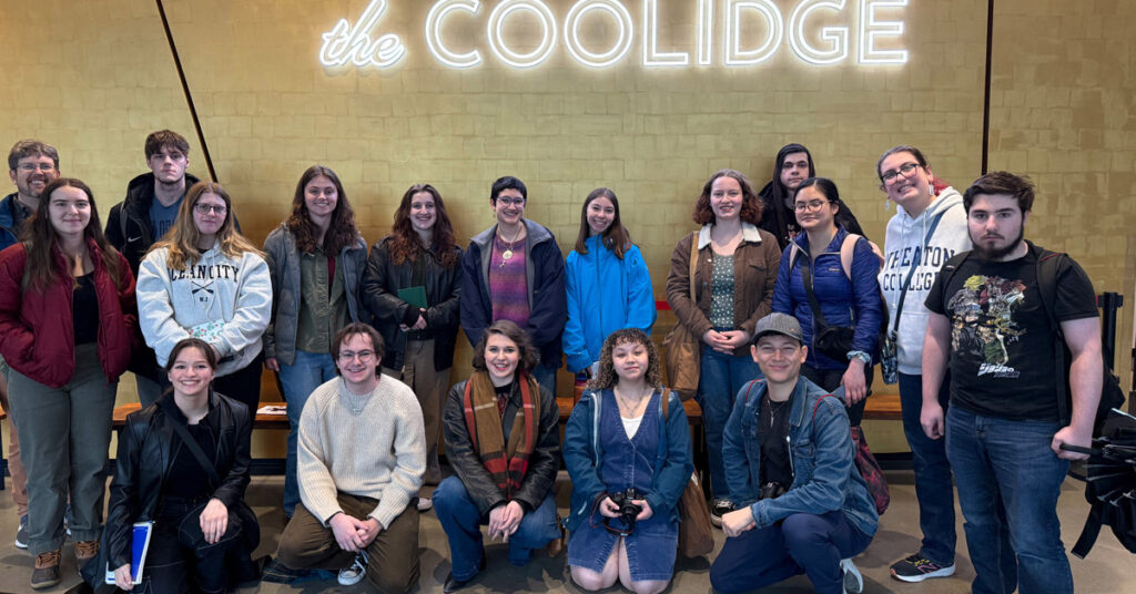A group of 18 young men and women sit or stand in front of a sign that reads the Coolidge.