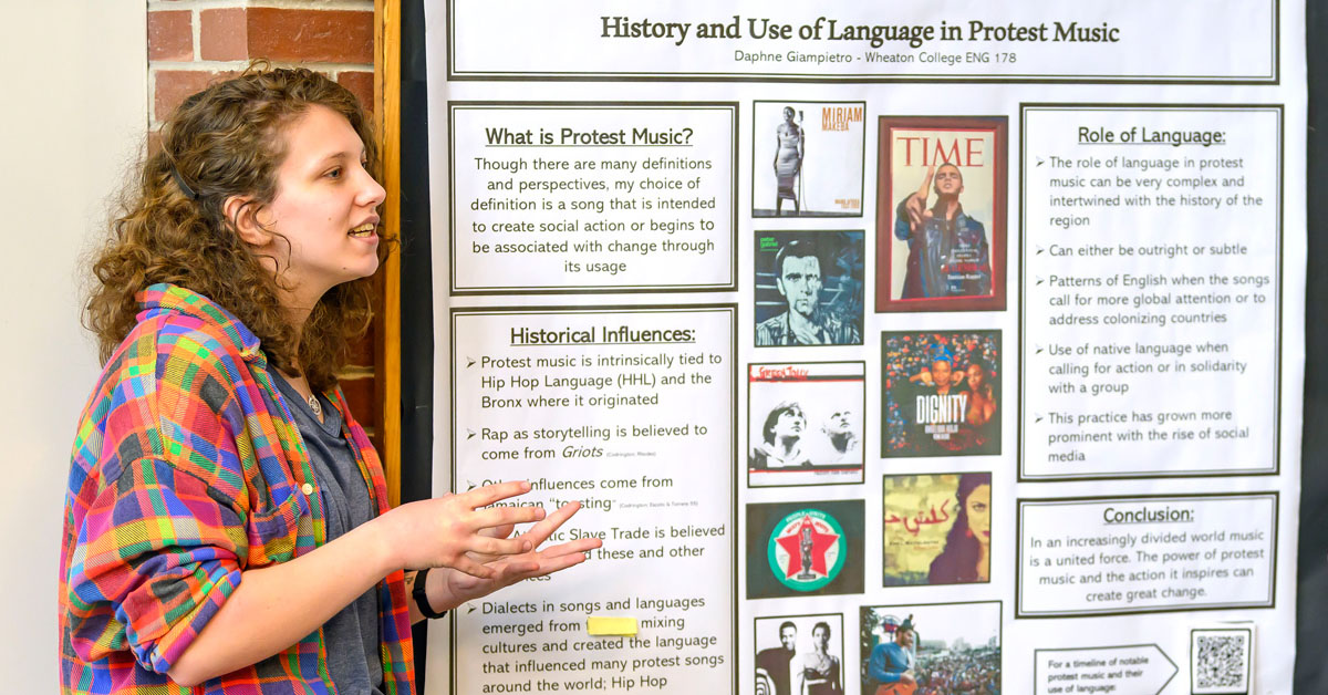 A young woman with wavy hair wearing a colorful shirt speaks while standing next to a poster titled Protest