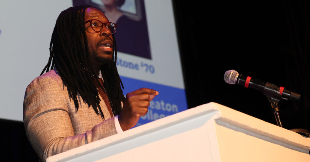 A Black man with long hair wearing glasses and a beige sportcoat speaks from a podium.