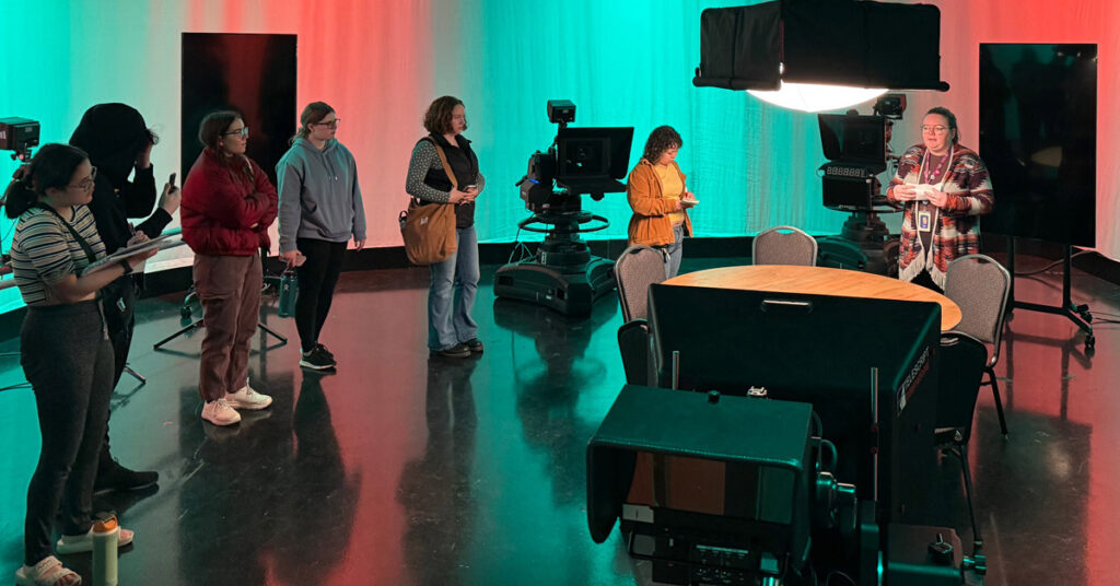 A woman speaks to a group of college-aged students in a television studio.